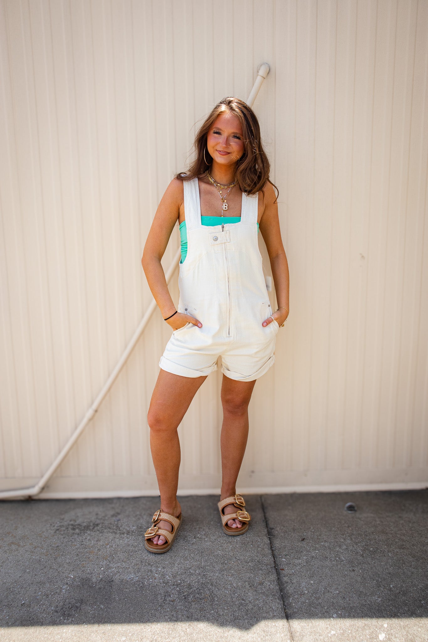 Model wearing a white denim romper with wide straps, front zipper detail, rolled hem shorts, and pockets, styled with a bright turquoise tube top, layered gold jewelry, and tan buckle sandals for a trendy summer boutique outfit.