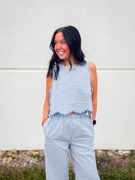 Blue and white gingham sleeveless top with scalloped hem and blue trim, featuring adjustable tie sides and a back button closure, styled with matching wide leg gingham pants for a coastal summer look.