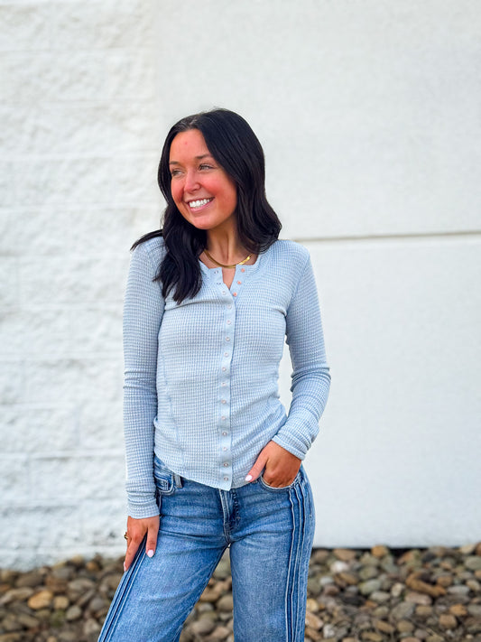 Woman wearing a light blue long-sleeve shirt and jeans standing against a white wall.