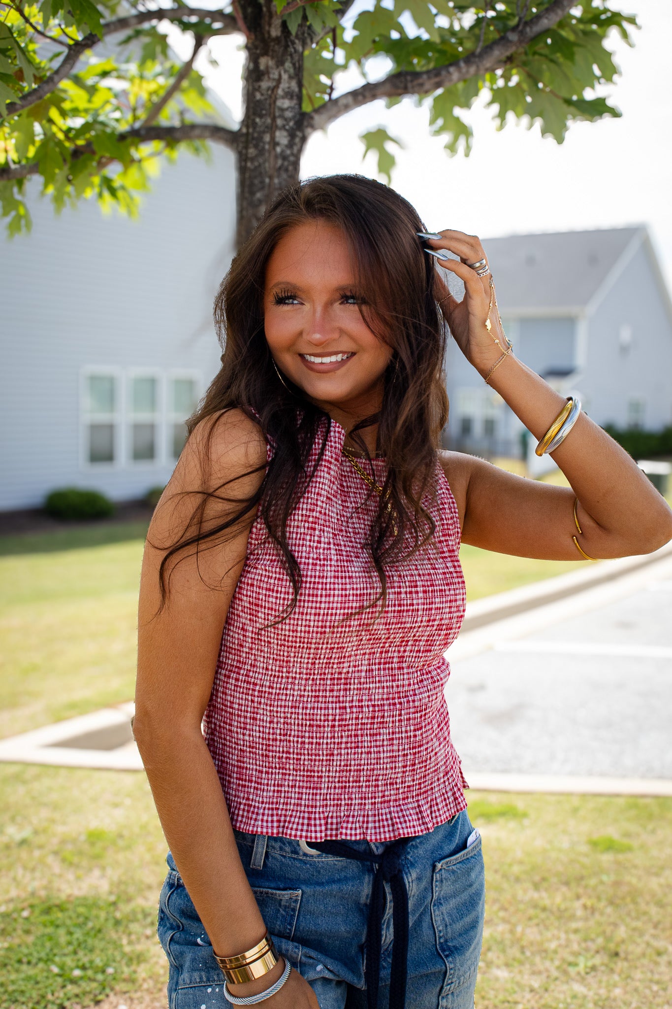 Model wearing a red gingham smocked sleeveless top with a ruffle hem, styled with denim jeans and gold jewelry for a trendy summer boutique outfit perfect for patriotic holidays, game day, picnics, and casual weekend plans.