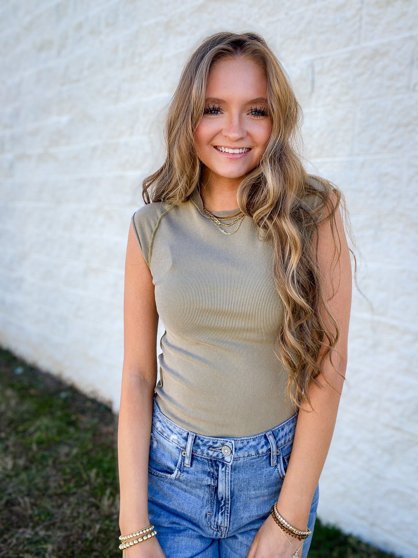 Woman wearing a green sleeveless top and blue jeans against a white wall.