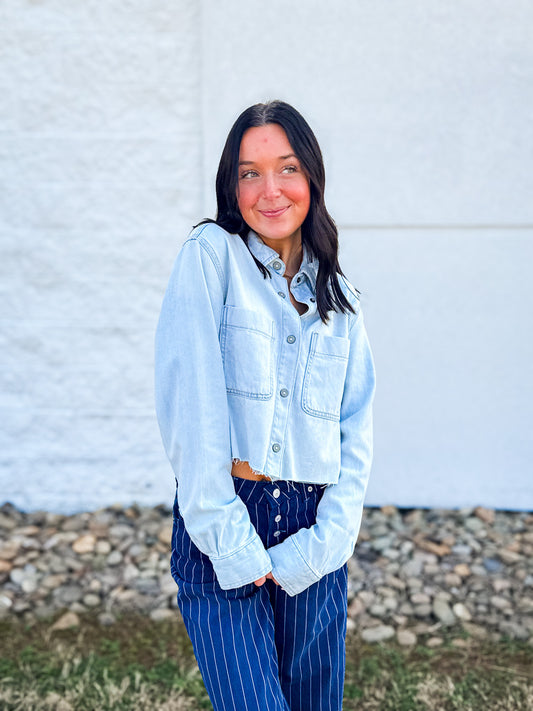 Woman wearing a light blue denim shirt and blue striped pants standing against a white wall.