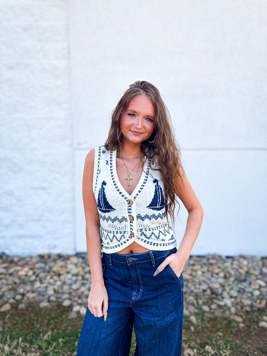 Woman wearing a sleeveless patterned top and blue jeans against a white wall.