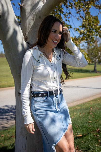 Woman in a white collared blouse and blue denim skirt standing next to a tree outdoors.