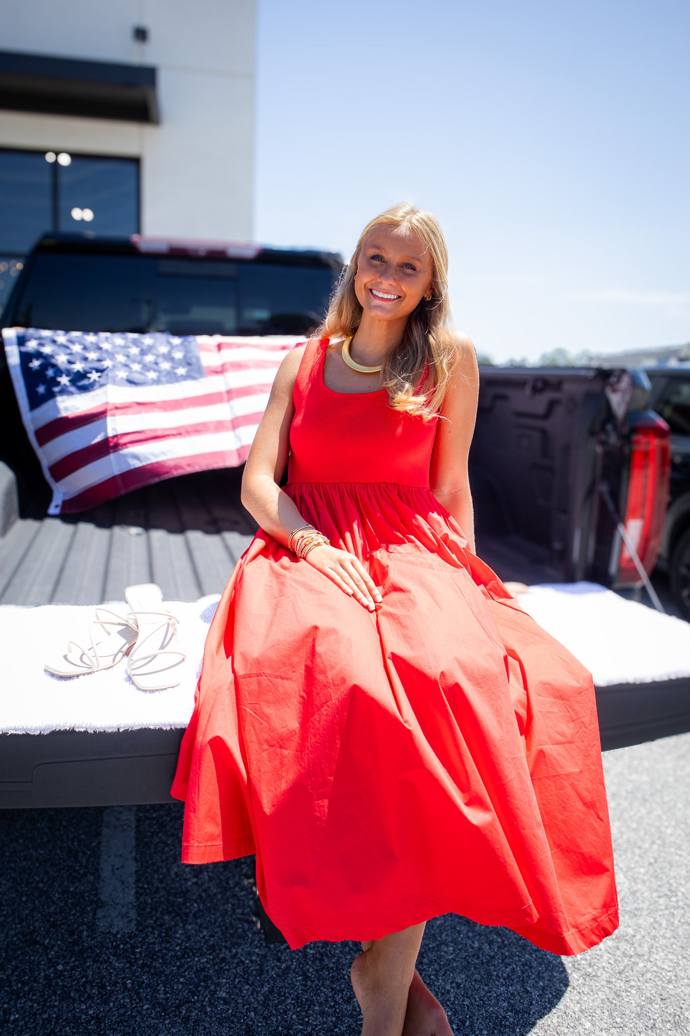 Model wearing a bright red sleeveless midi dress with a fitted tank-style bodice and flowy gathered skirt, styled with gold jewelry beside an American flag for a standout patriotic summer boutique outfit perfect for Memorial Day, Fourth of July, vacations, and warm-weather events.