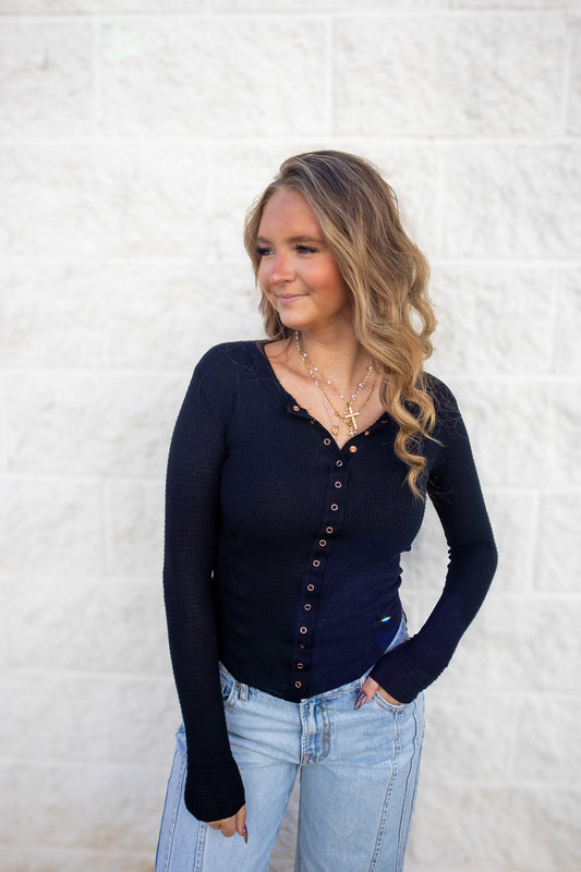 Woman wearing a black long-sleeve top with snap buttons against a white textured wall.