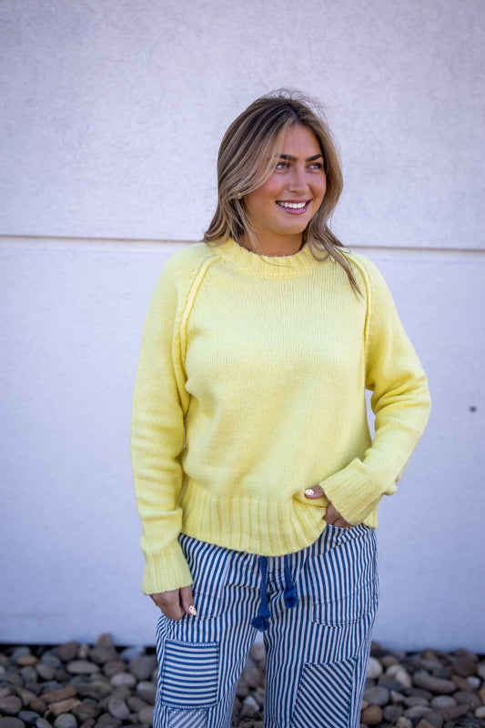 Woman wearing a yellow sweater and striped pants standing against a white wall.