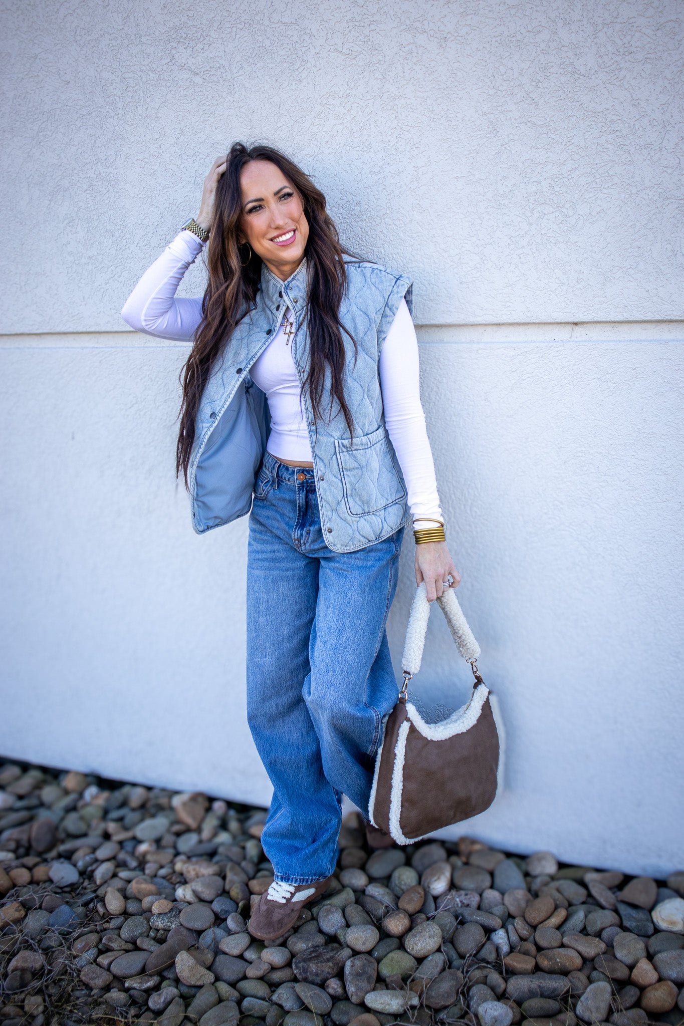 Woman wearing a denim vest and jeans, holding a brown bag against a white wall.