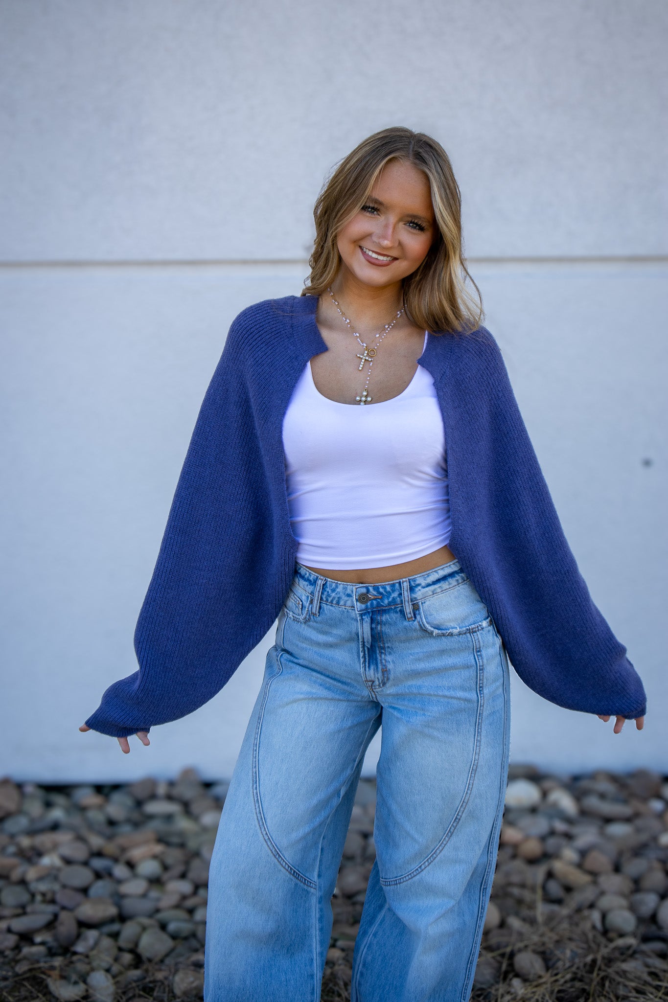 Woman wearing a blue cardigan, white top, and jeans against a light gray wall.