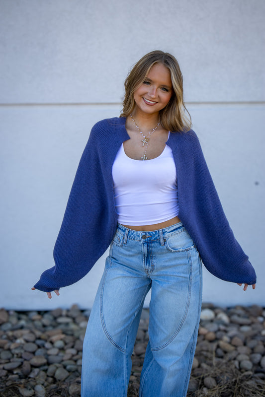 Woman wearing a blue cardigan, white top, and jeans against a light gray wall.