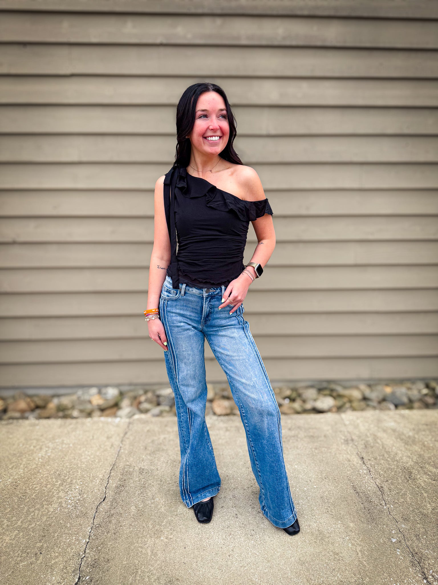 Woman wearing a black one-shoulder top and blue jeans standing against a beige wall.