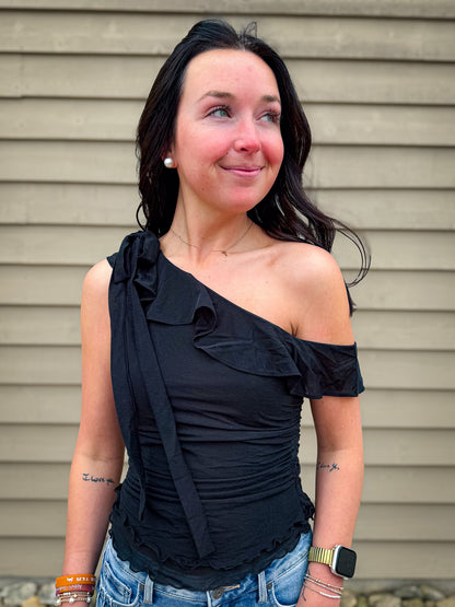 Woman wearing a black one-shoulder top with ruffles against a beige wall.