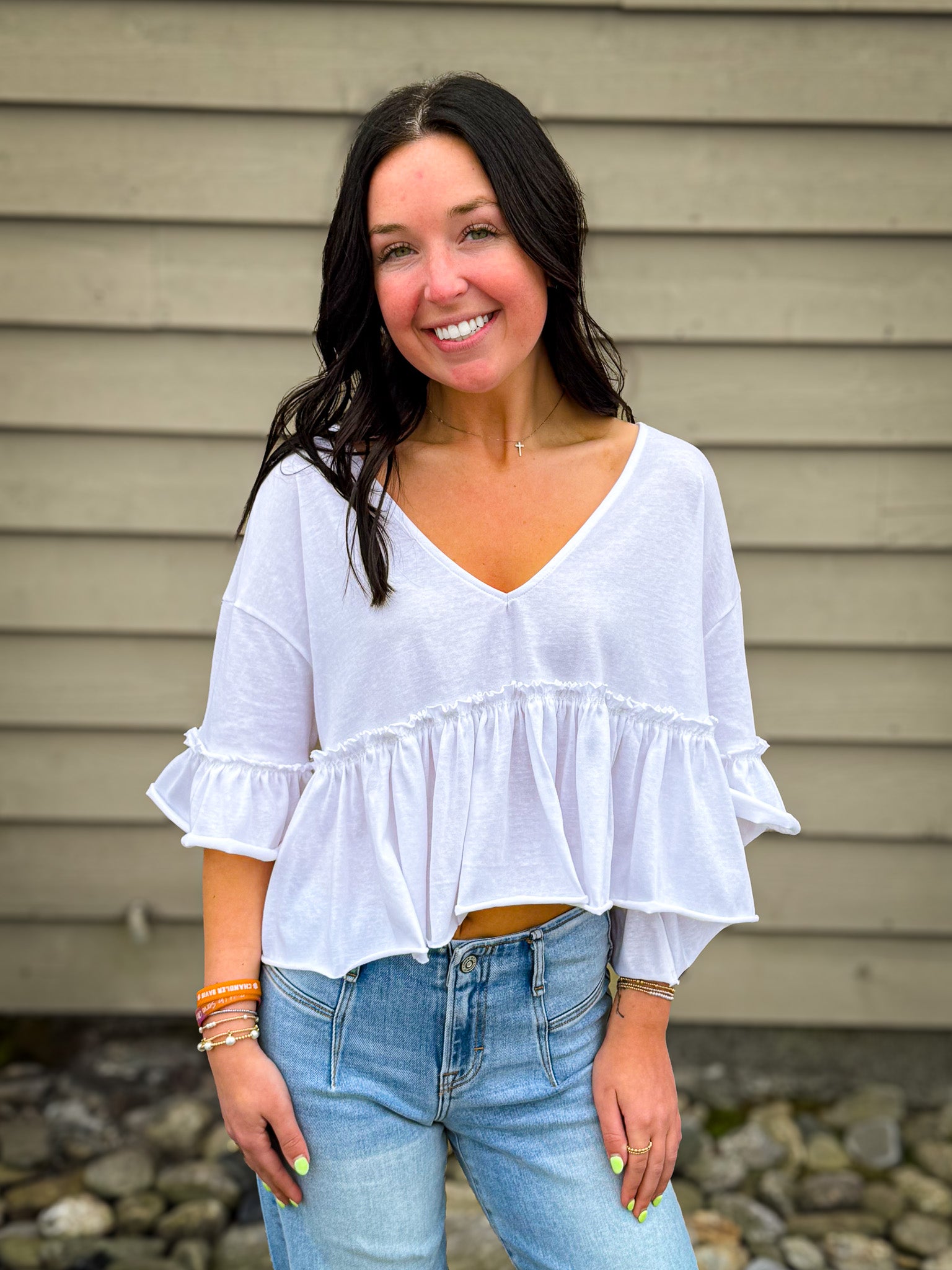 Woman wearing a white blouse with ruffled sleeves and blue jeans standing against a beige wall.