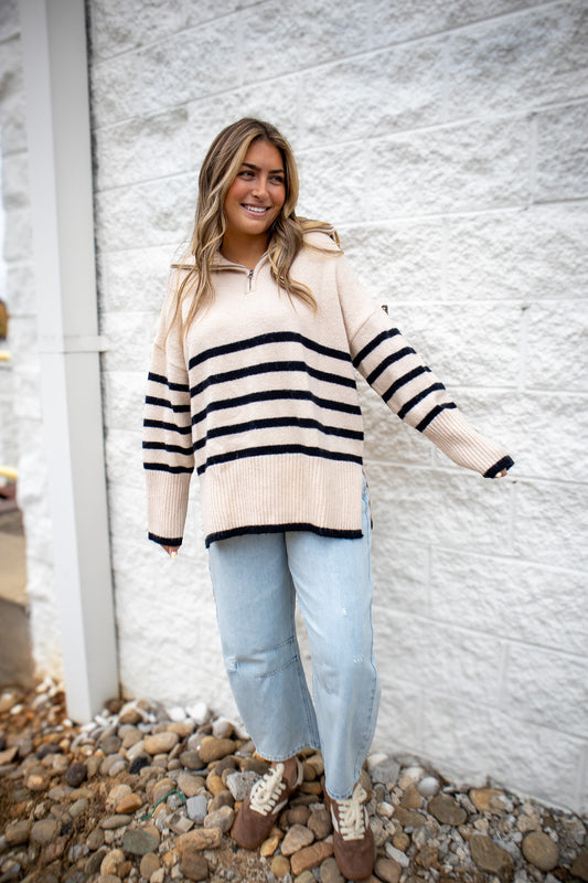 Woman wearing a striped sweater and light blue jeans standing against a white wall.