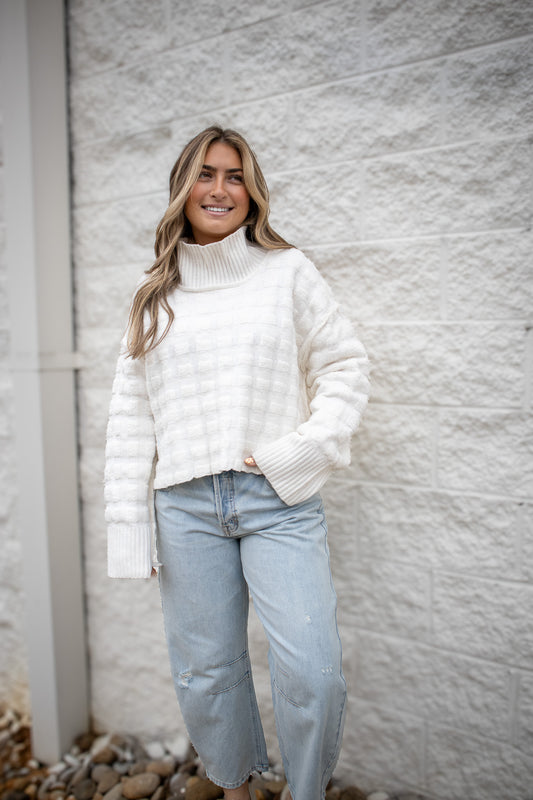 Woman wearing a white sweater and light blue jeans standing against a textured gray wall.