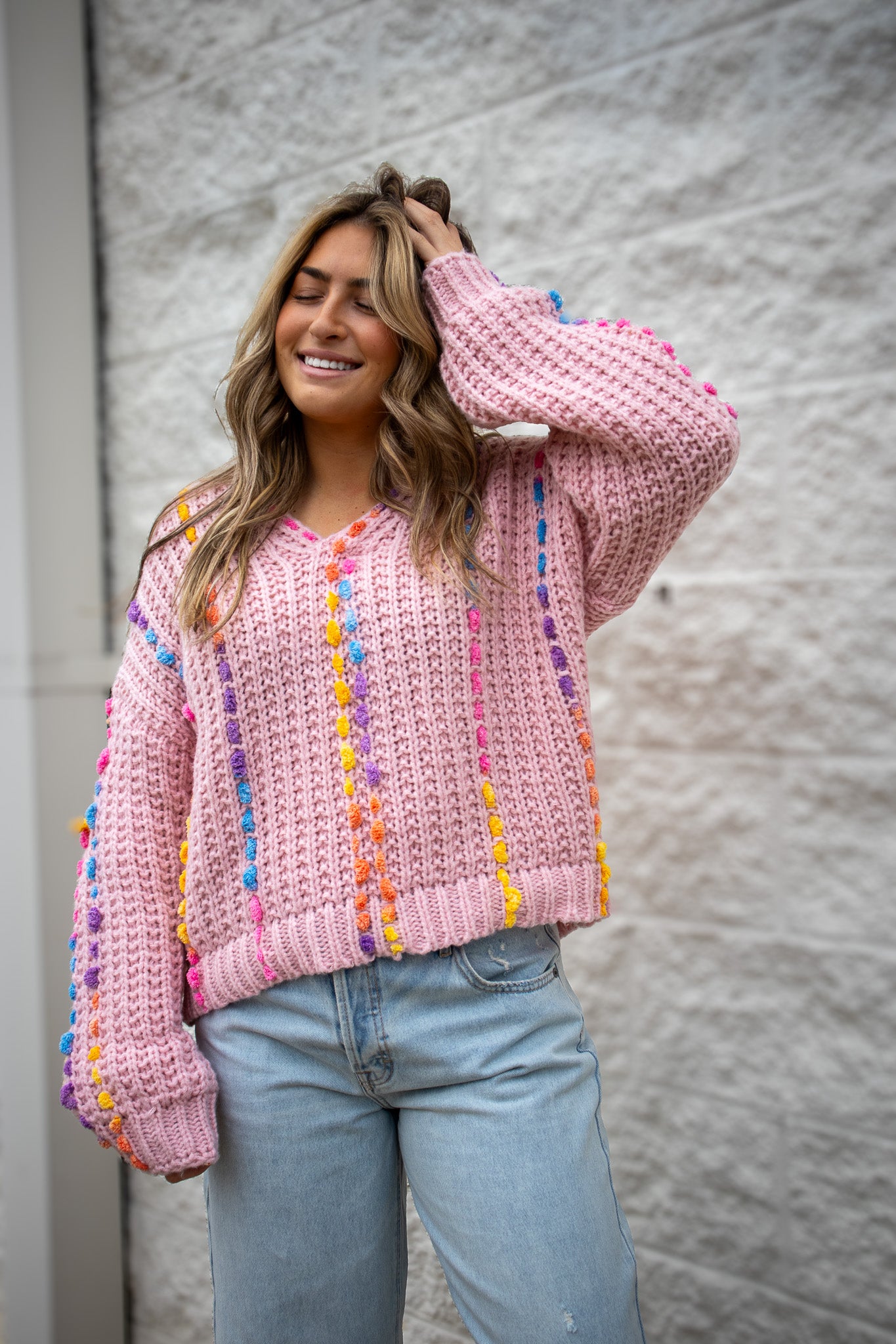 Woman wearing a pink sweater with colorful pom-poms against a gray wall.