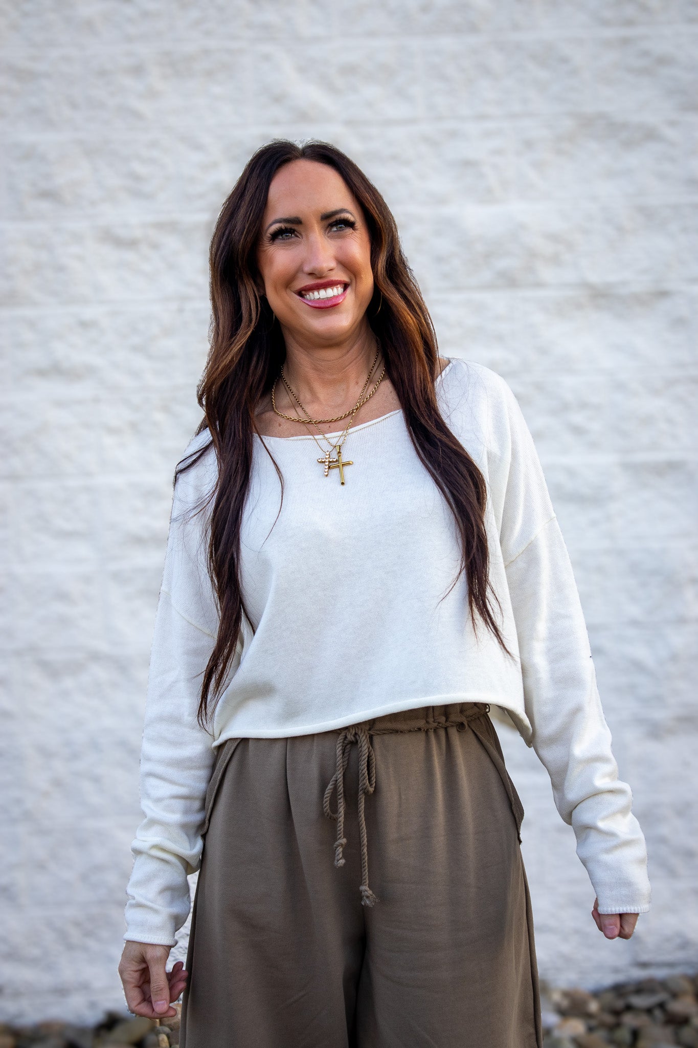 Woman wearing a white top and brown pants against a light gray wall.