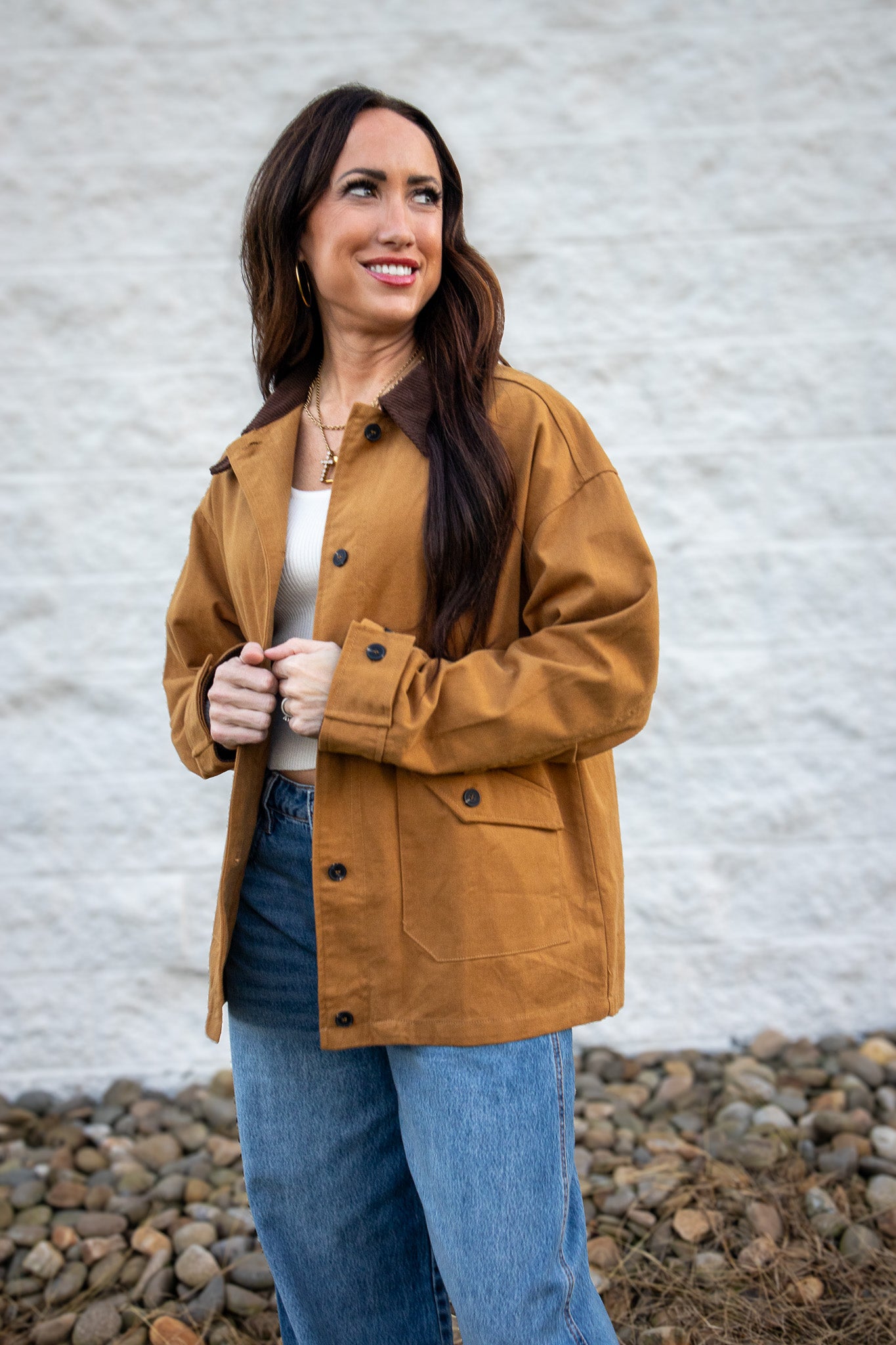 Woman wearing a brown jacket and blue jeans standing against a white wall.