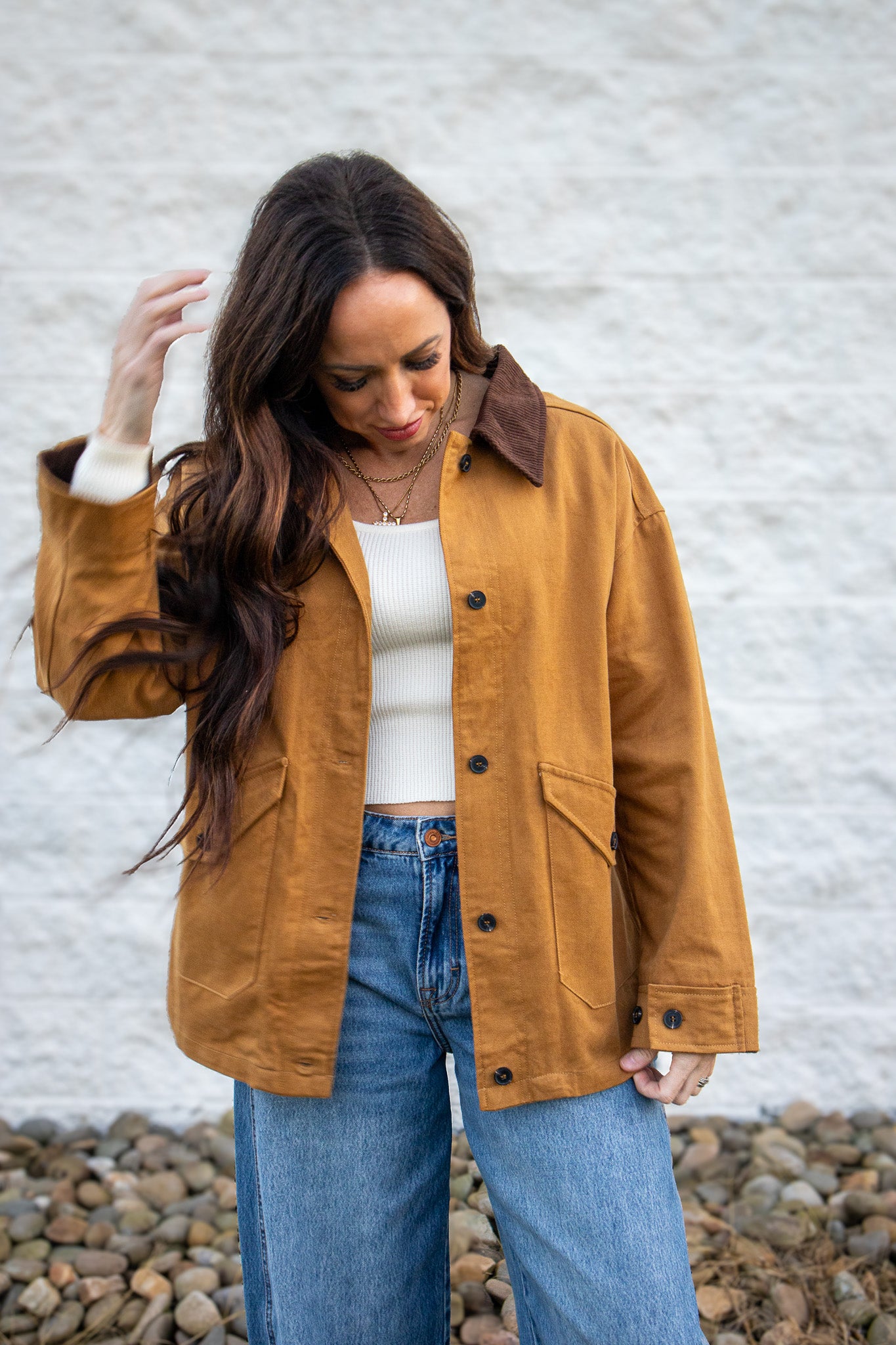 Woman wearing a tan jacket and blue jeans against a white wall.