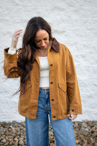 Woman wearing a tan jacket and blue jeans against a white wall.