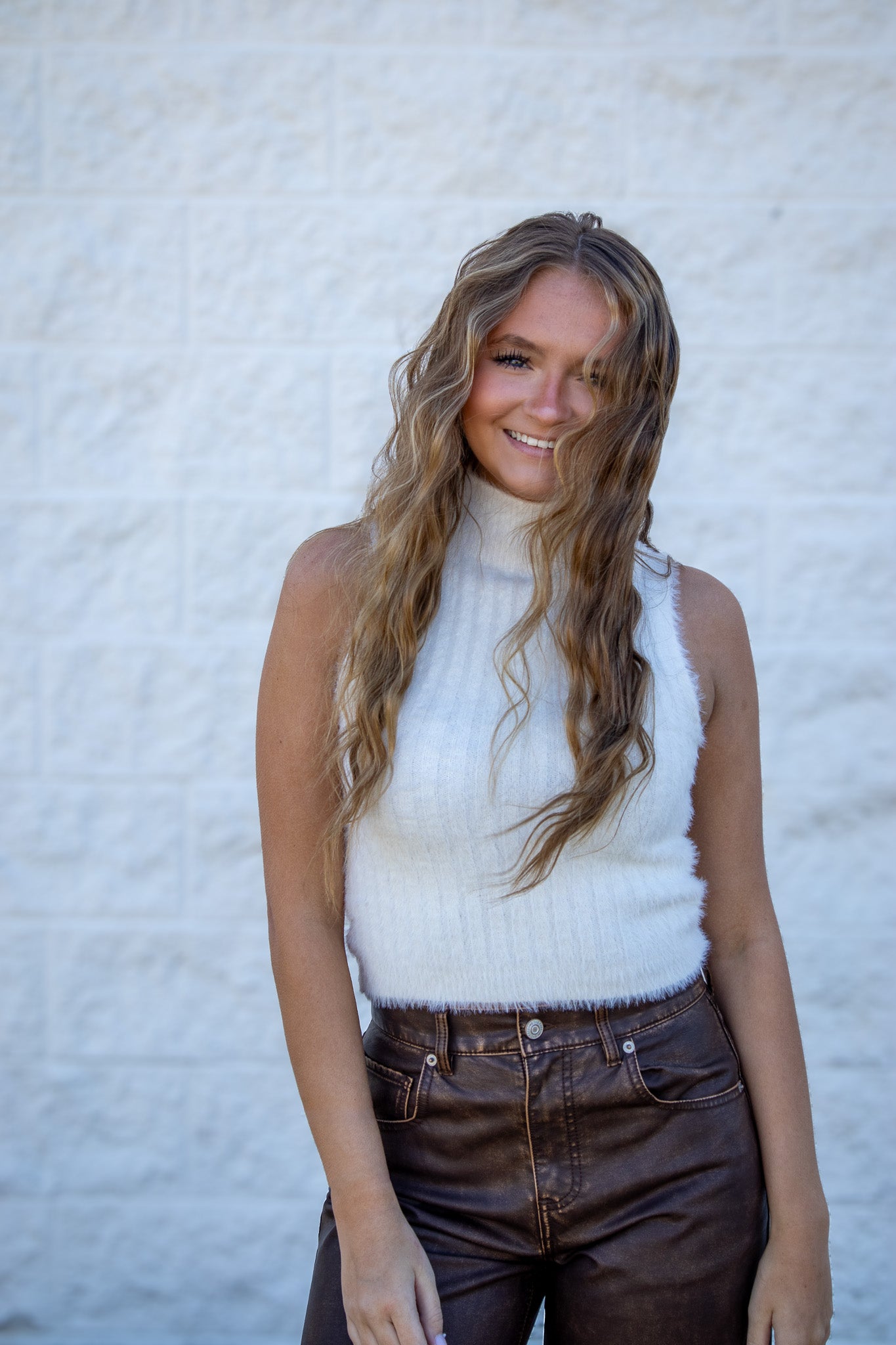 Woman wearing a white sleeveless sweater and brown pants against a light textured wall.