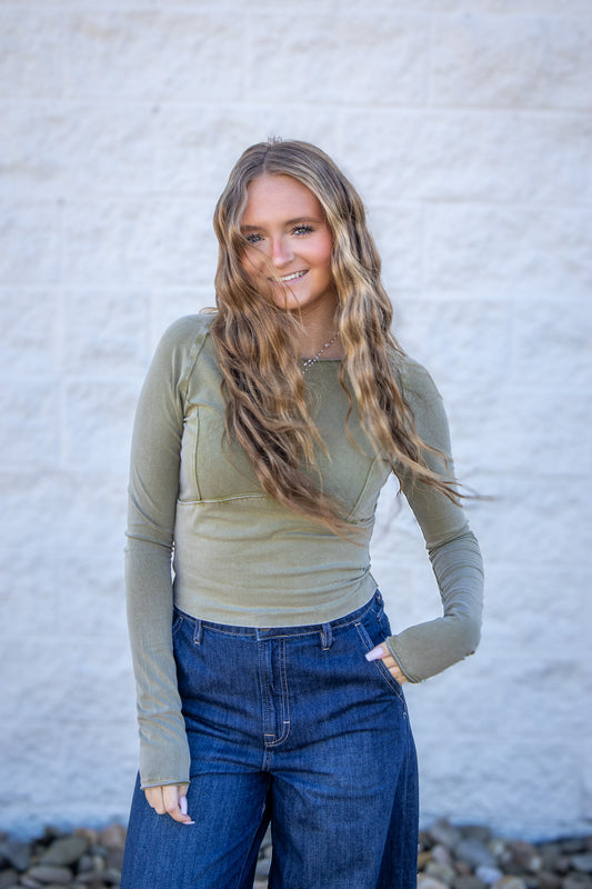 Woman wearing a green long-sleeve top and blue jeans against a white wall.