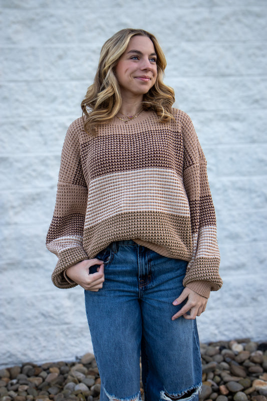 Woman wearing a brown and beige sweater with blue jeans against a gray wall.