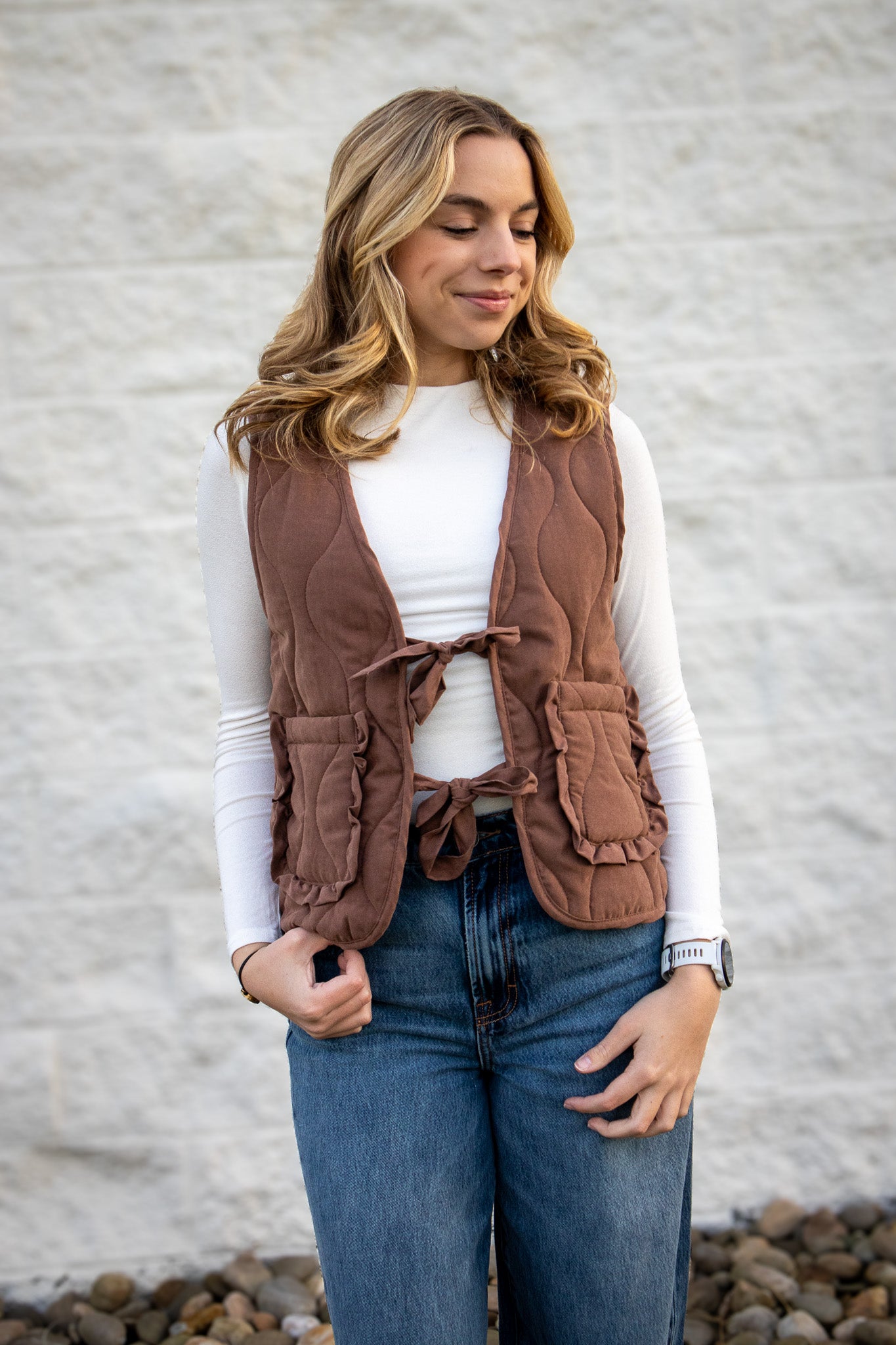 Woman wearing a brown vest over a white shirt and blue jeans against a gray wall.