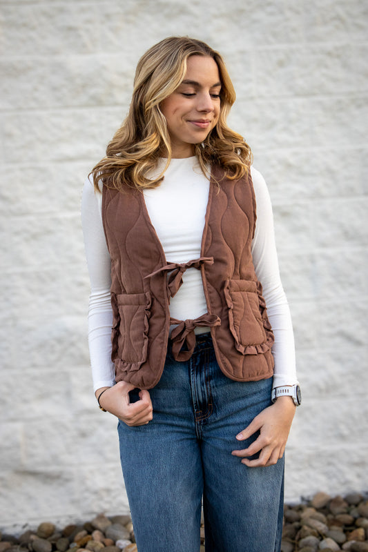 Woman wearing a brown vest over a white shirt and blue jeans against a gray wall.