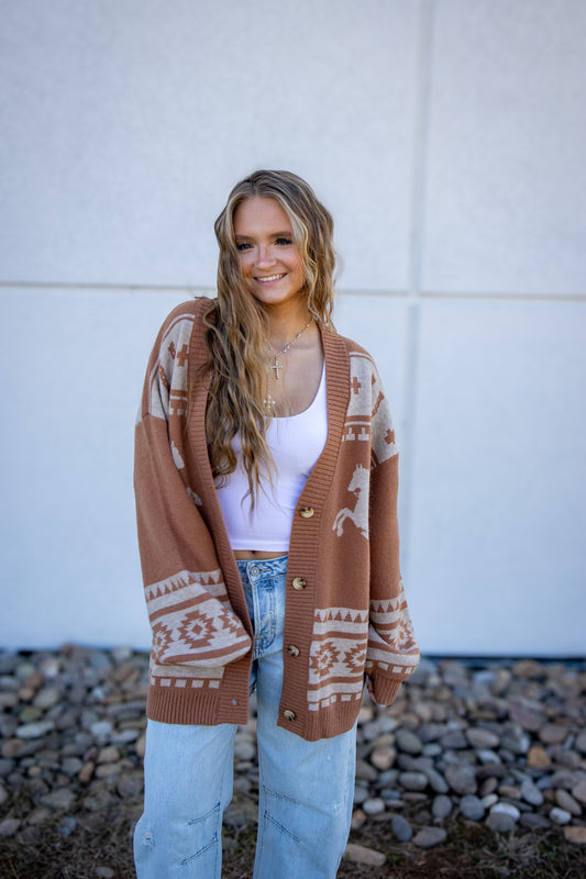 Woman wearing a patterned brown cardigan over a white top and light blue jeans, standing against a white wall with a rocky ground.