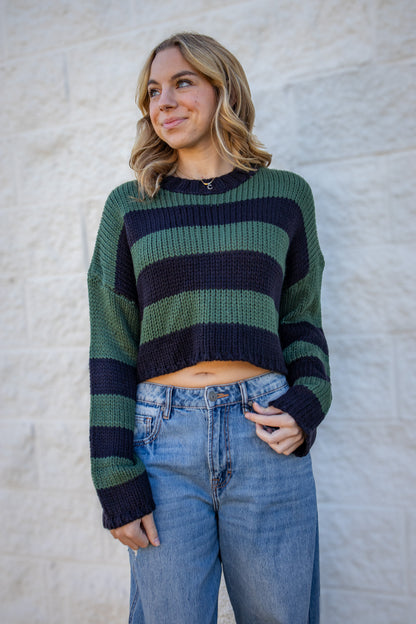 Woman wearing a green and navy striped sweater with blue jeans against a textured white wall.