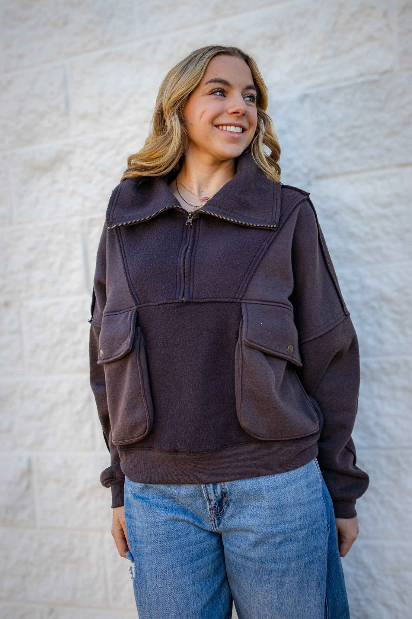 Woman wearing a brown pullover with pocket details against a light stone wall.