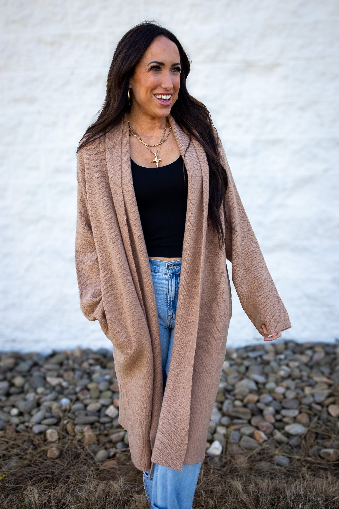 Woman wearing a long beige cardigan over a black top and light blue jeans, standing against a white wall.