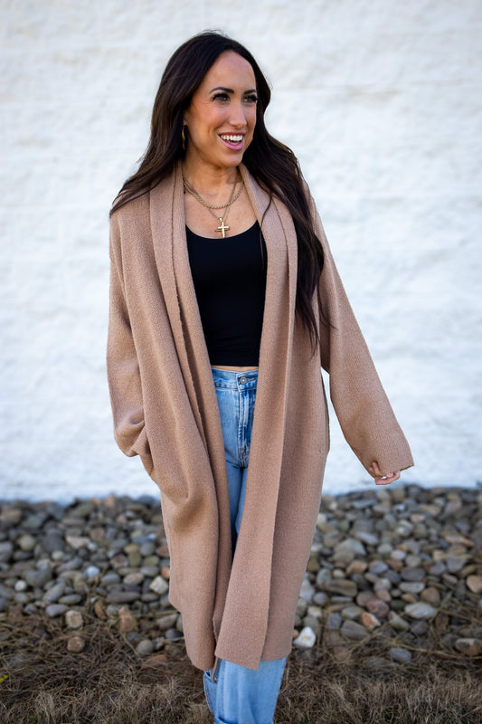 Woman wearing a long beige cardigan over a black top and light blue jeans, standing against a white wall.