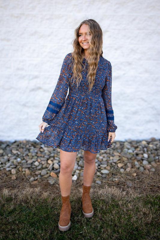 Woman wearing a blue floral dress standing against a white wall with a stone base.