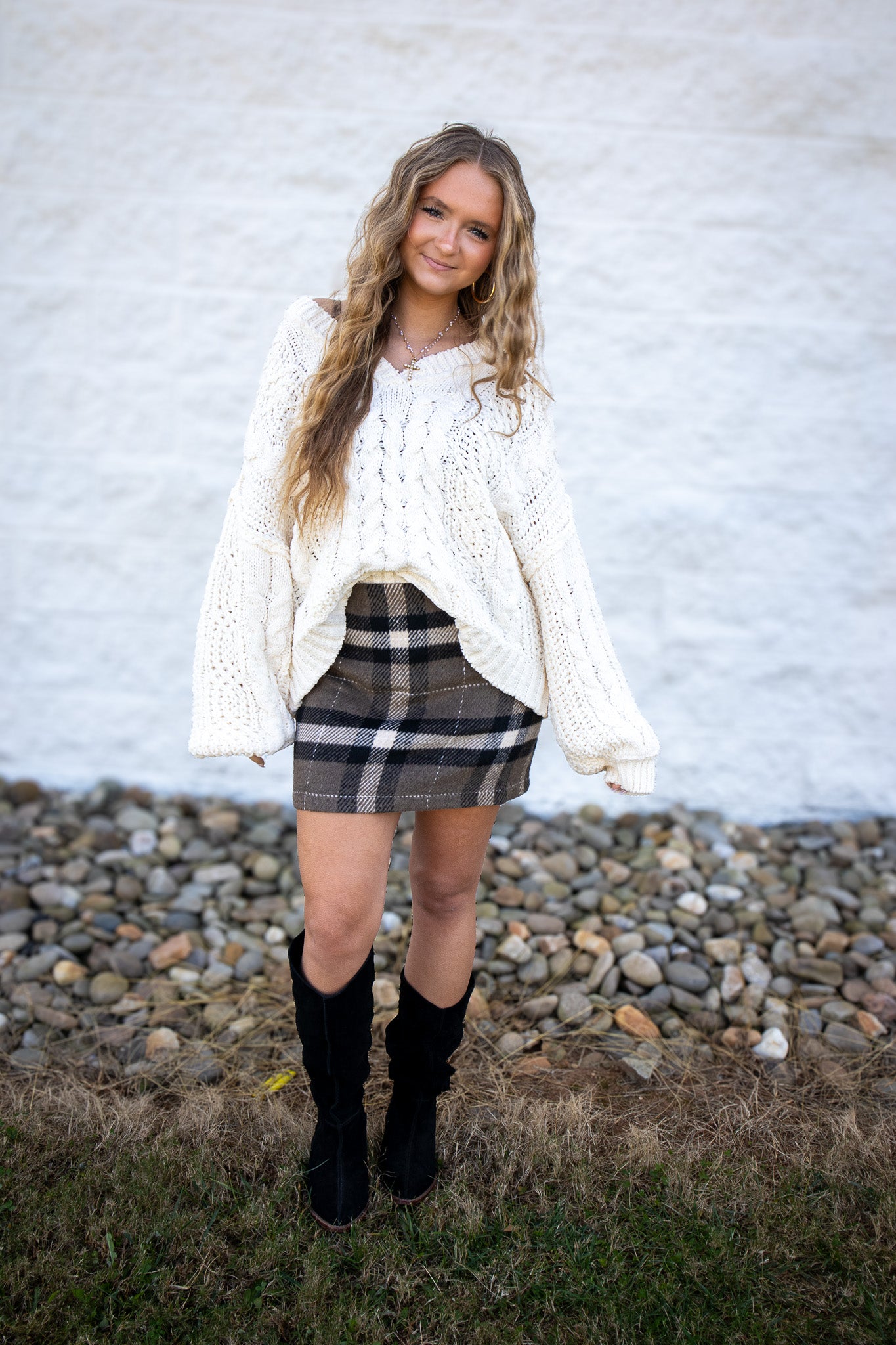 Woman wearing a white sweater, plaid skirt, and black boots against a white brick wall.