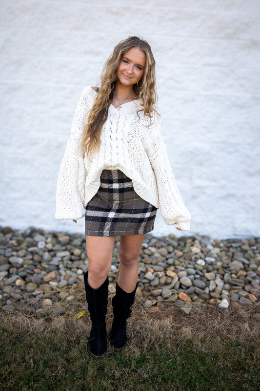 Woman wearing a white sweater, plaid skirt, and black boots against a white brick wall.