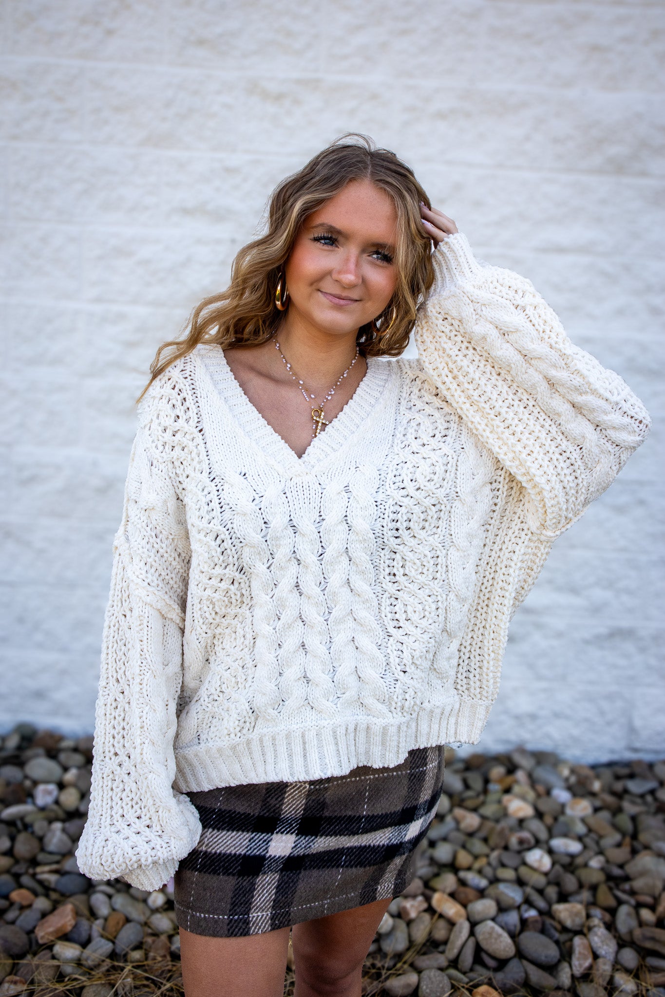 Woman wearing a white chunky knit sweater and plaid skirt against a textured wall.