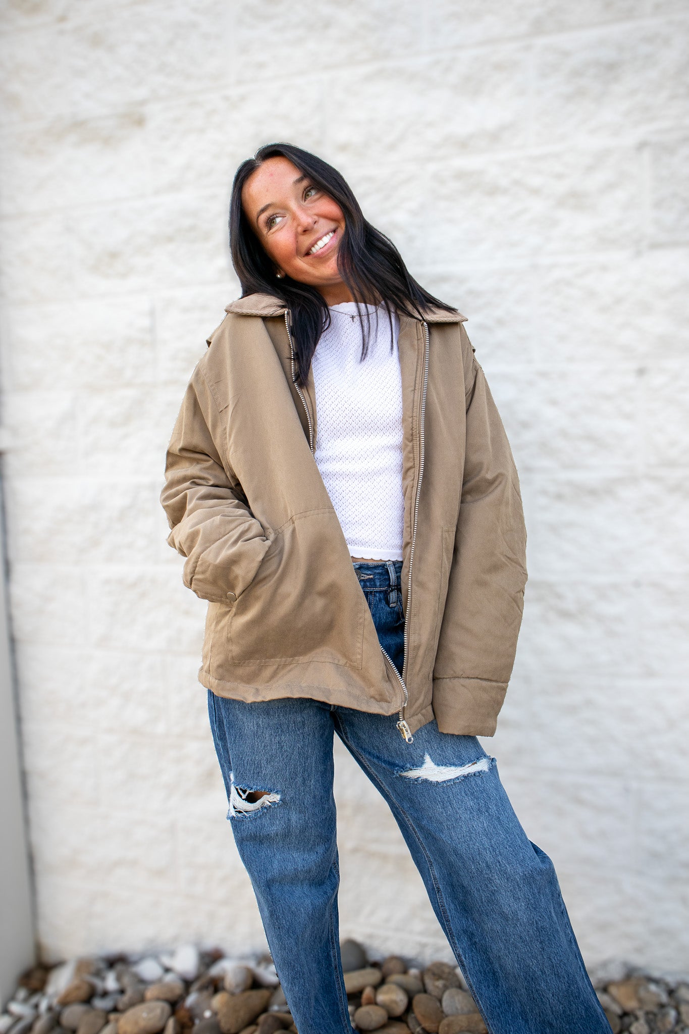 Woman wearing a beige jacket, white shirt, and blue jeans against a textured wall.