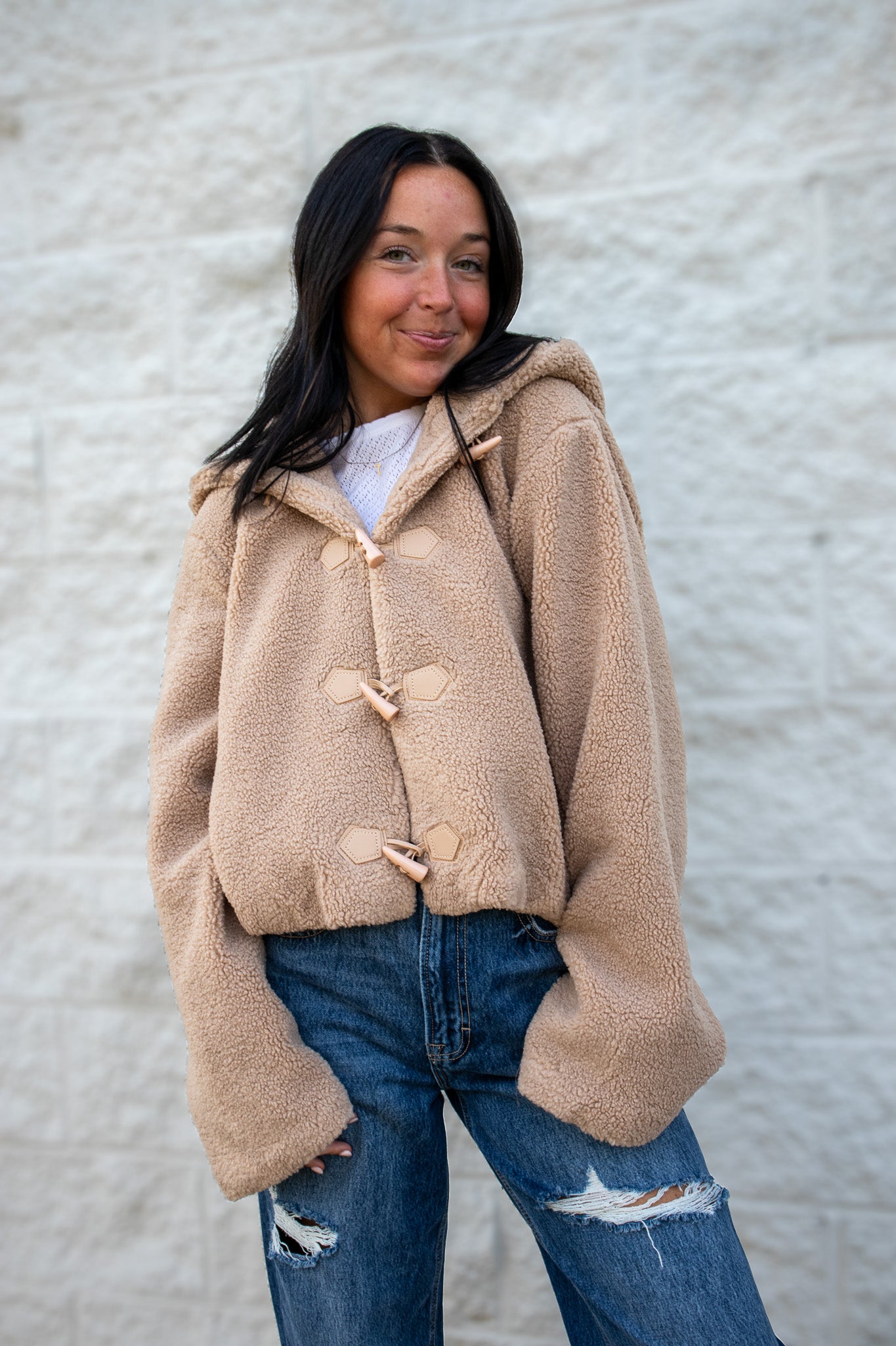 Woman wearing a beige fleece jacket with button details against a textured white wall.