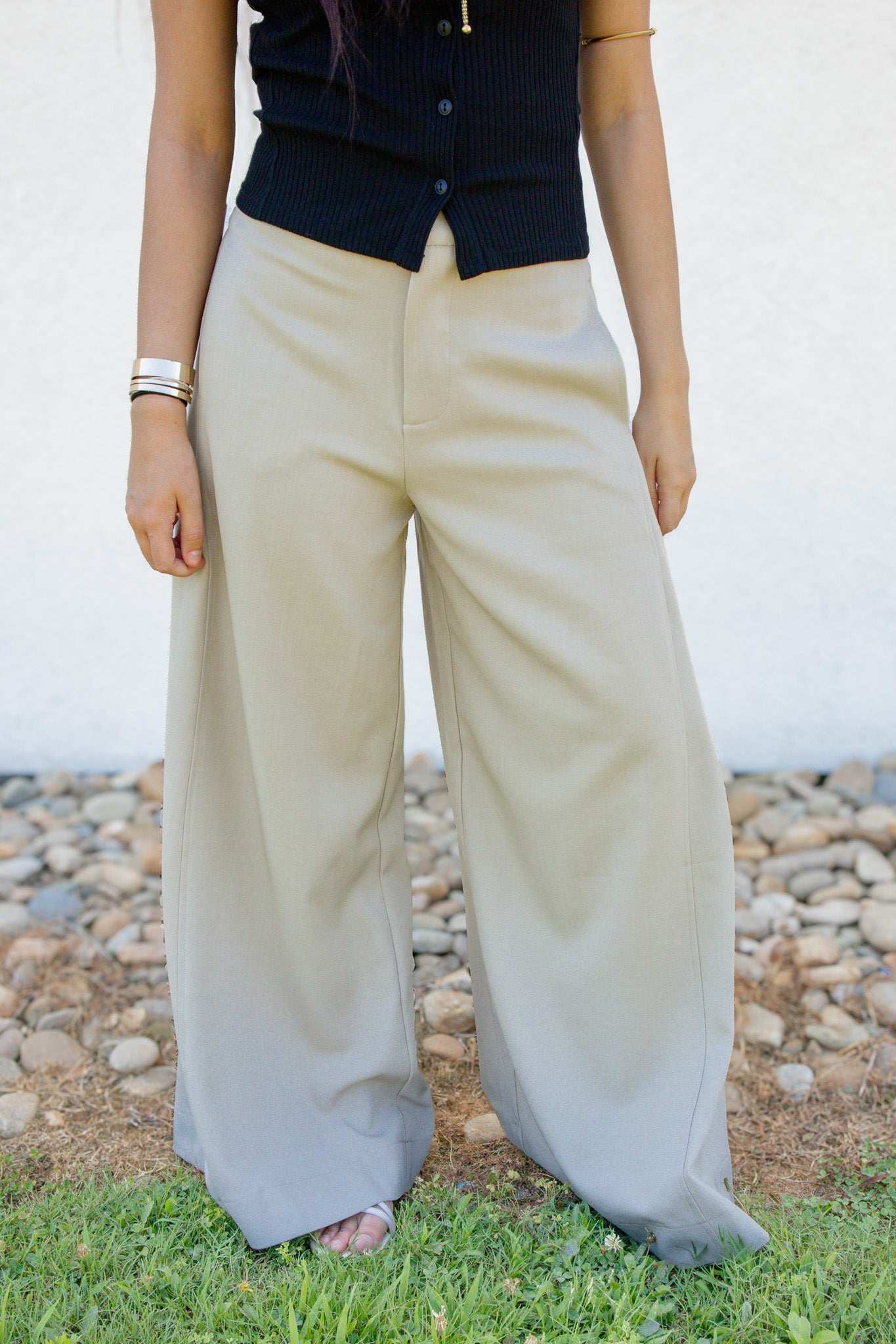 A person wearing wide-leg beige pants and a black ribbed top, standing outdoors on grass with a white wall and pebble ground in the background.