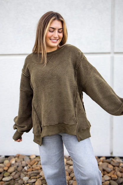 Woman wearing an olive green sweater and light blue jeans standing against a white wall with pebbles at the bottom.