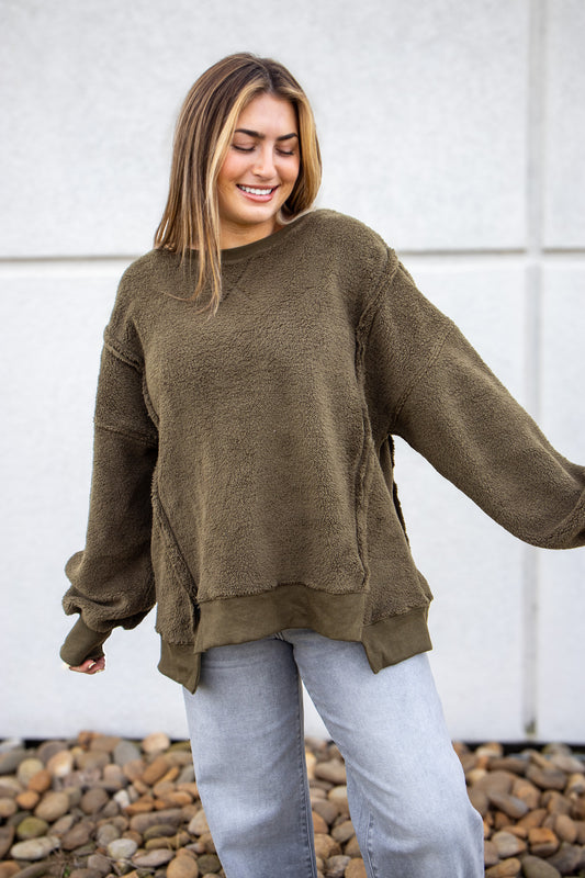 Woman wearing an olive green sweater and light blue jeans standing against a white wall with pebbles at the bottom.