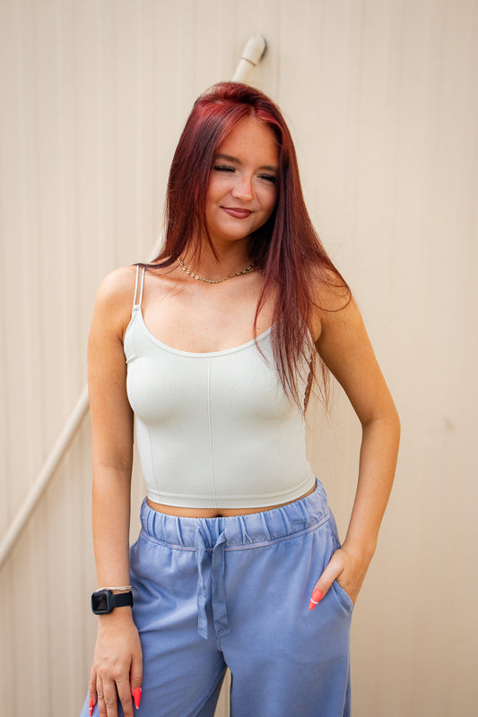 Woman wearing a light-colored tank top and blue pants against a beige wall.