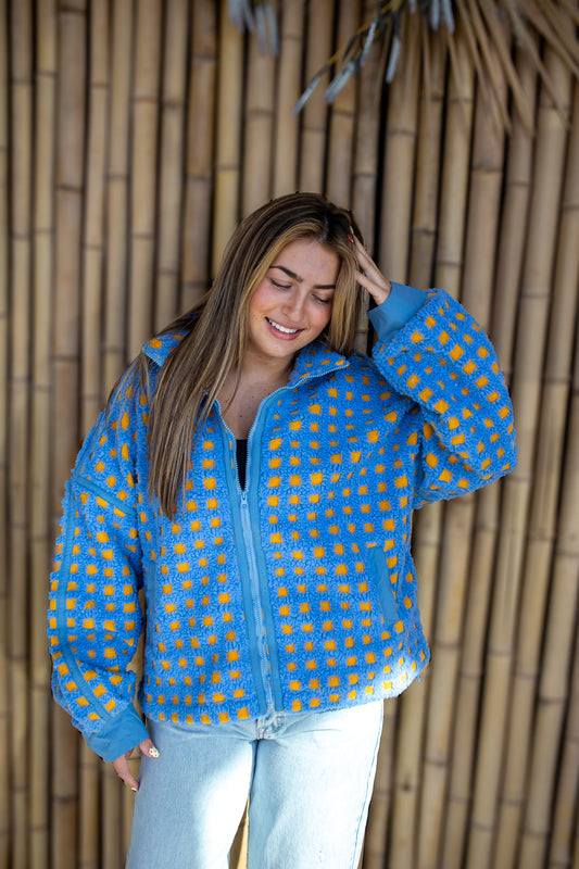 Woman wearing a blue and yellow patterned jacket against a wooden wall.