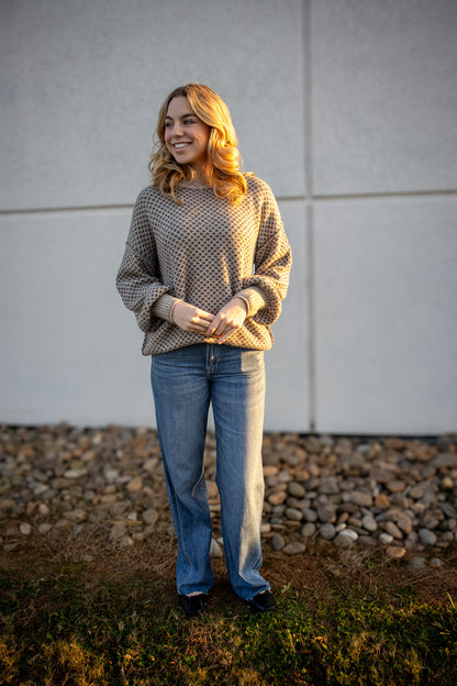 Woman wearing a patterned sweater and jeans standing outdoors against a plain wall.