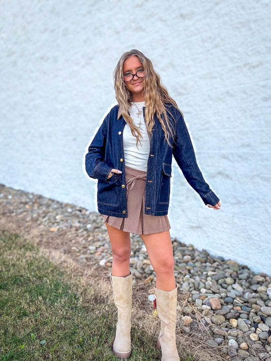 Person wearing a denim jacket, white shirt, brown skirt, and beige boots standing against a textured wall.