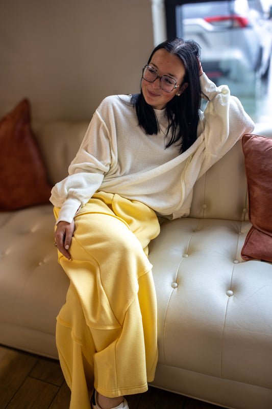 Person wearing a white sweater and yellow pants sitting on a beige couch with red pillows.