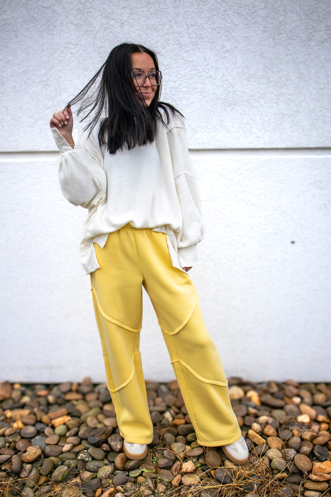 Person wearing a white sweater and yellow pants standing on pebbles with a white wall background