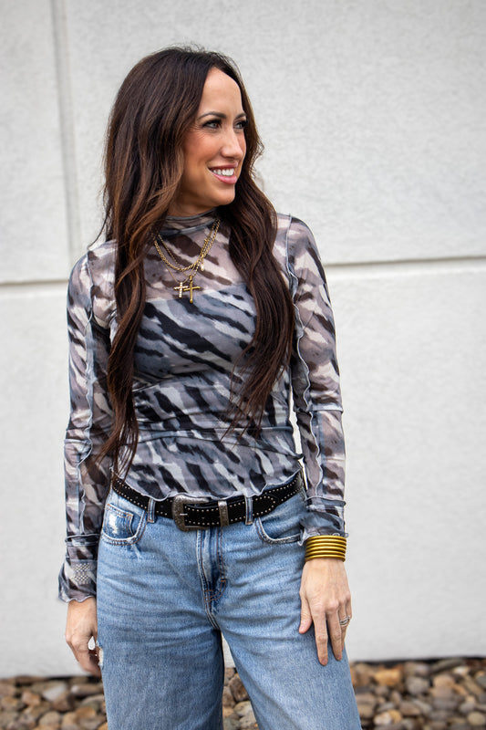 Woman wearing a patterned long-sleeve top and jeans against a gray wall.
